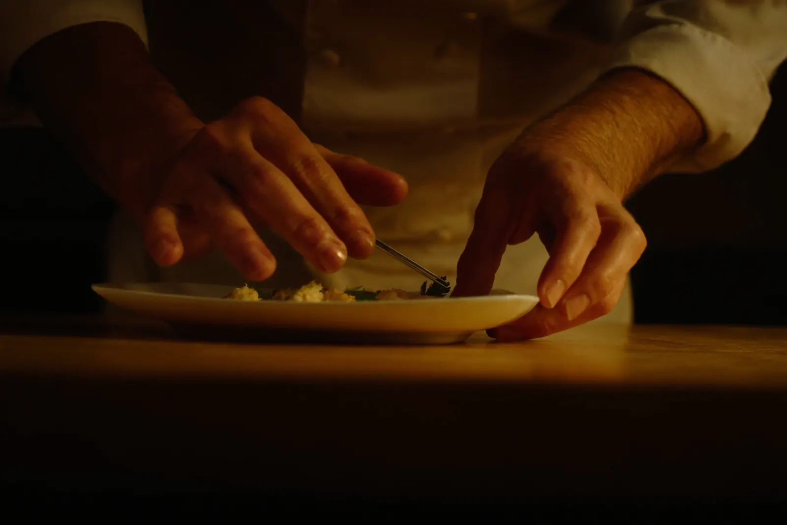 Mani di uno chef che completano con cura un piatto, in una cucina professionale dalla luce calda.