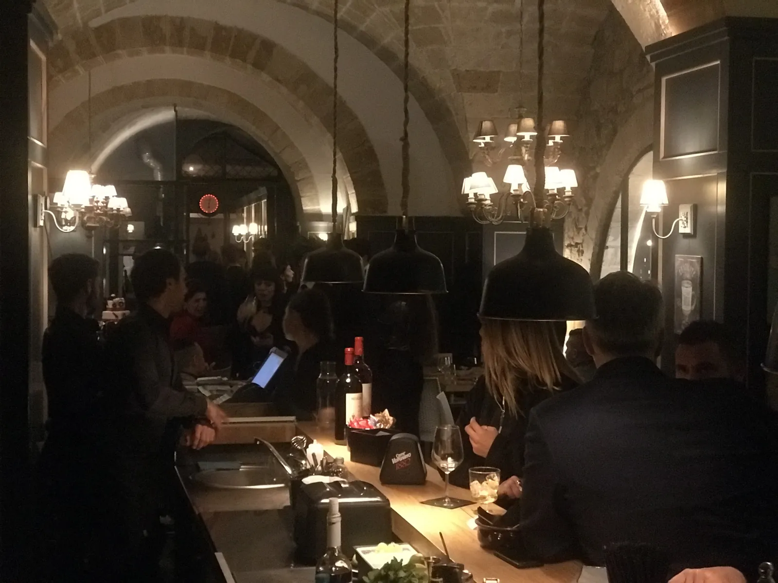 Bancone bar affollato la sera, sotto volte in pietra e lampadari accesi.