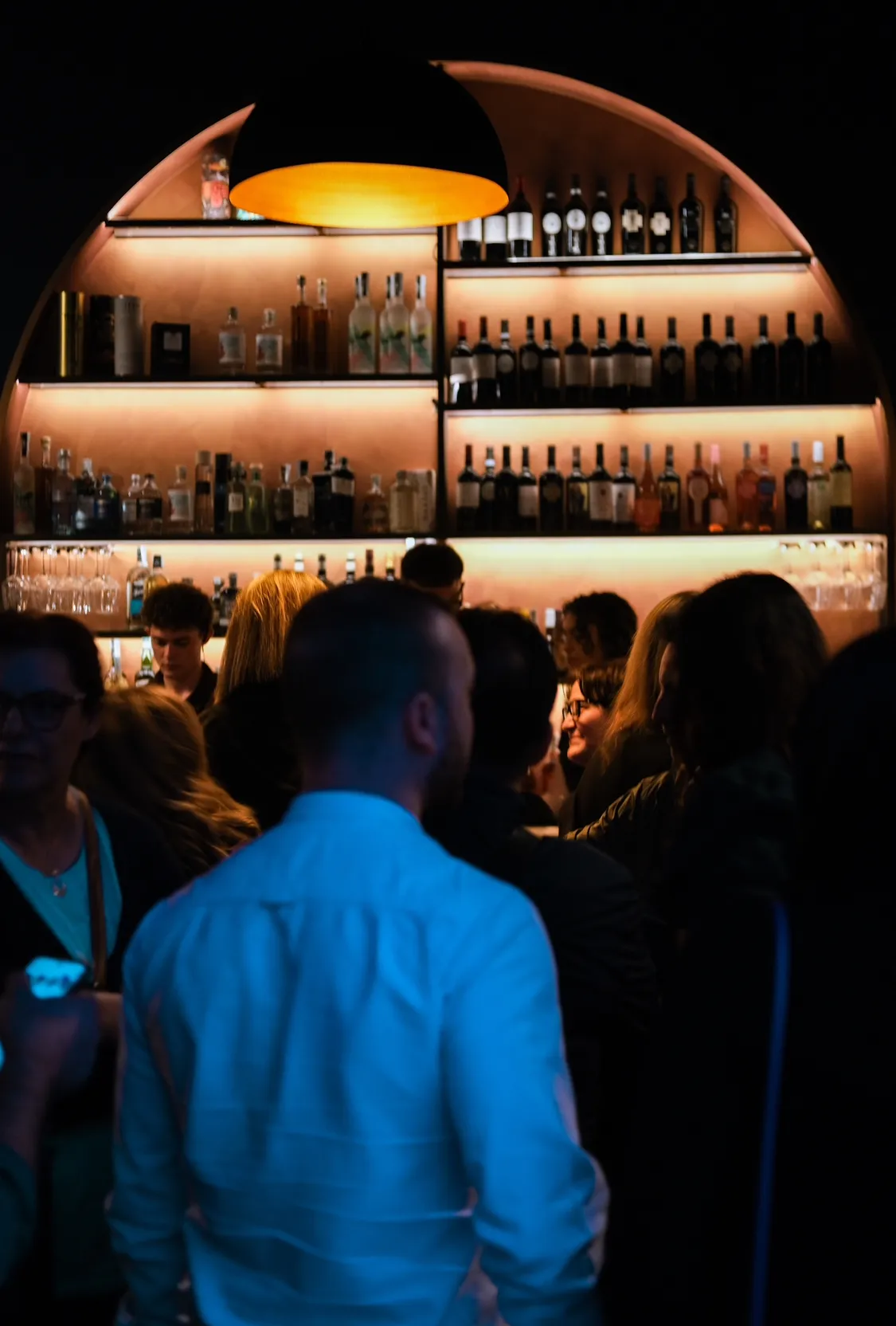 Serata al banco bar: ospiti davanti allo scaffale ad arco illuminato.