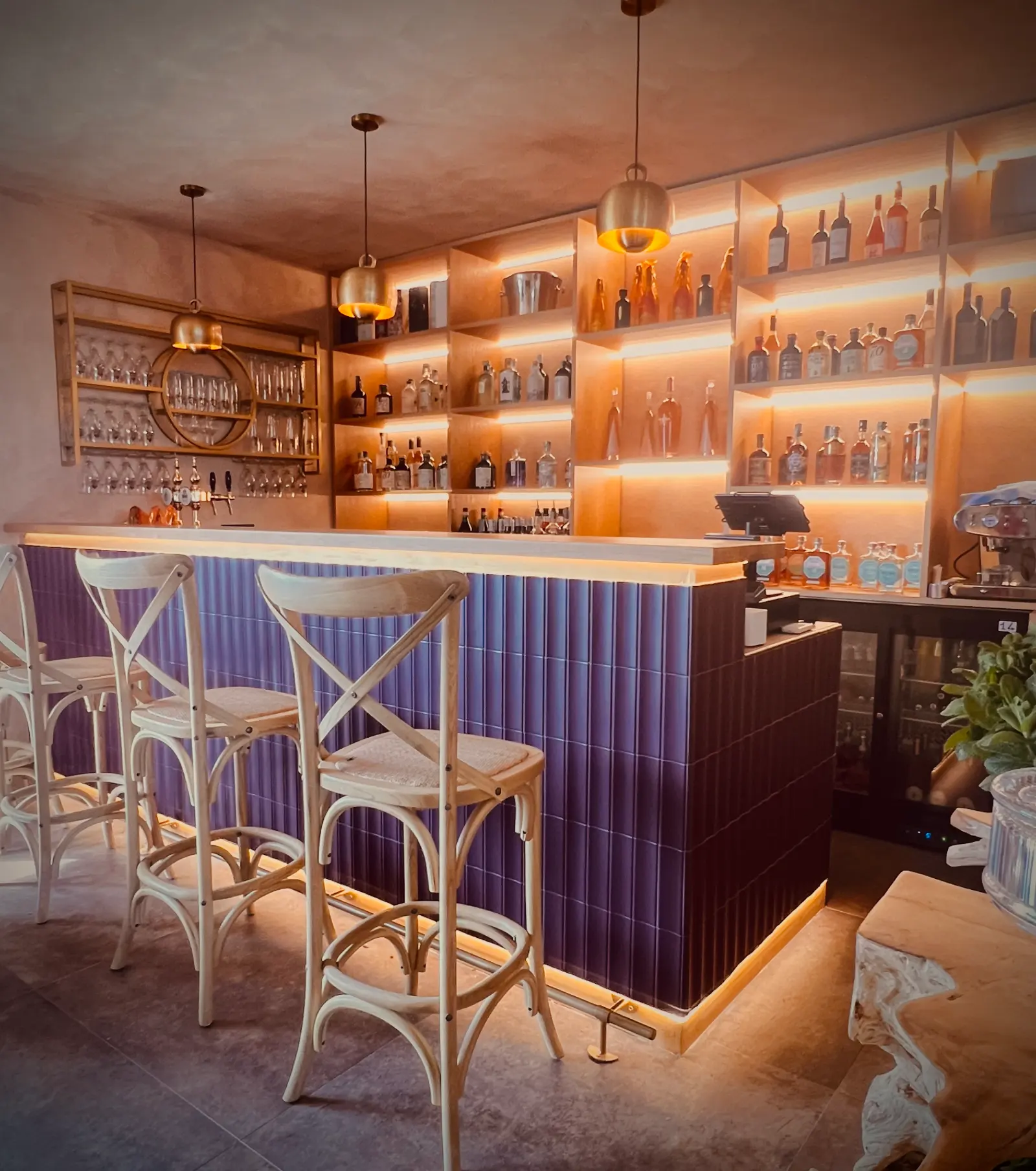 Cocktail bar con bancone rivestito di piastrelle viola, sgabelli in legno chiaro e mensole illuminate.
