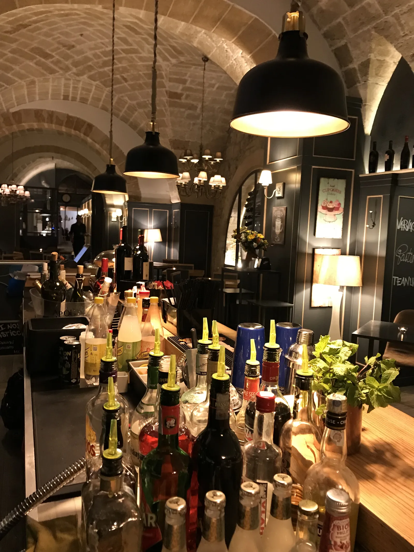 Bancone bar con bottiglie in primo piano e lampade industriali sotto le volte in pietra.