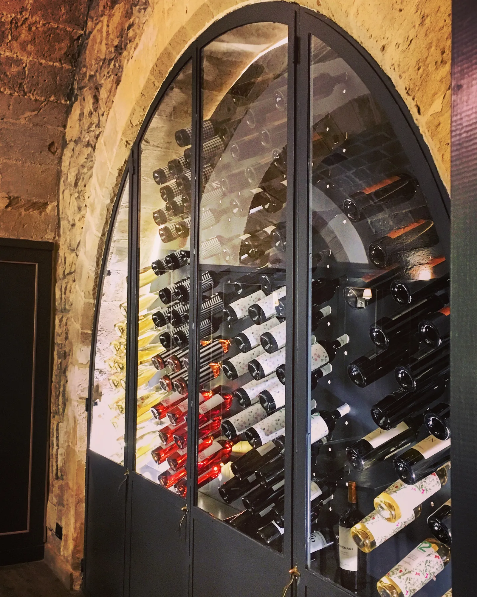 Cantina a vista con bottiglie coricate in una vetrina ad arco, contro un muro in pietra.