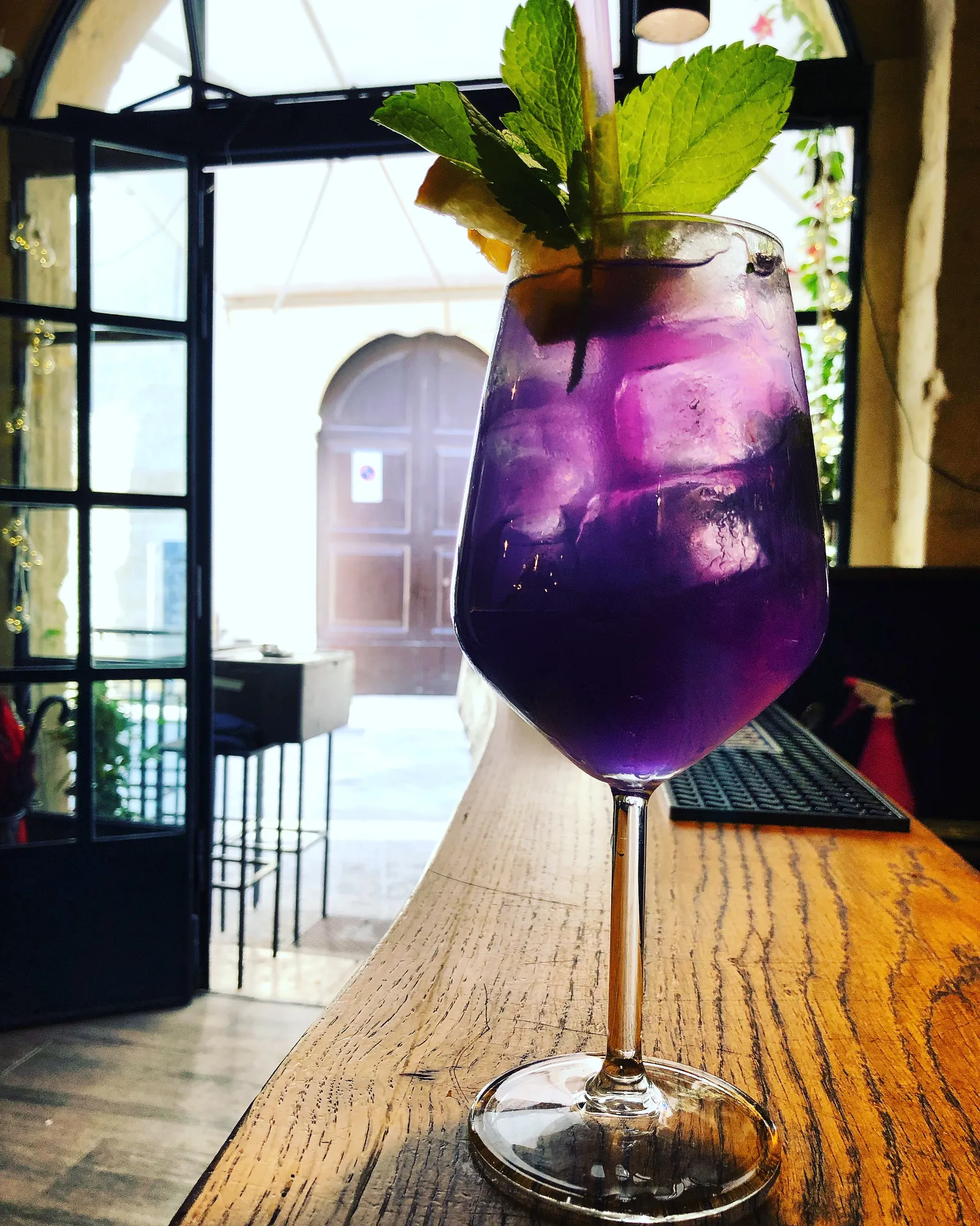 Cocktail viola con menta in un calice, sul bancone in legno accanto a una porta vetrata.
