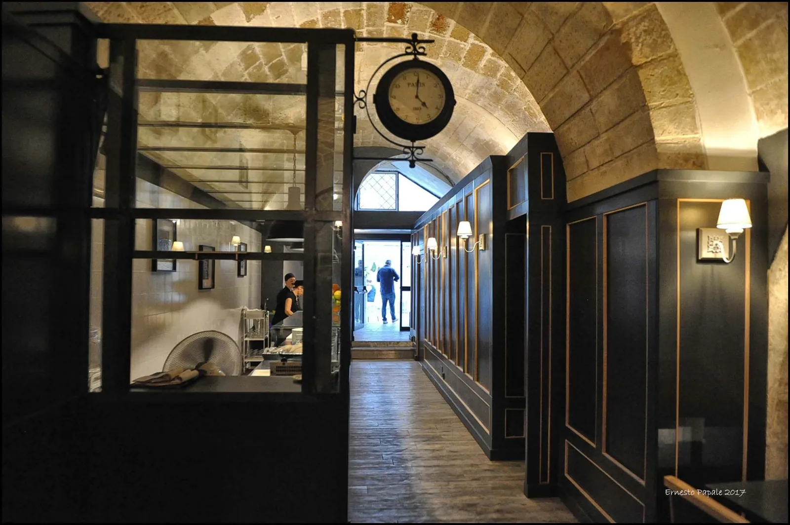 Corridoio con volta in pietra, boiserie scura e un orologio da stazione sopra la porta vetrata.