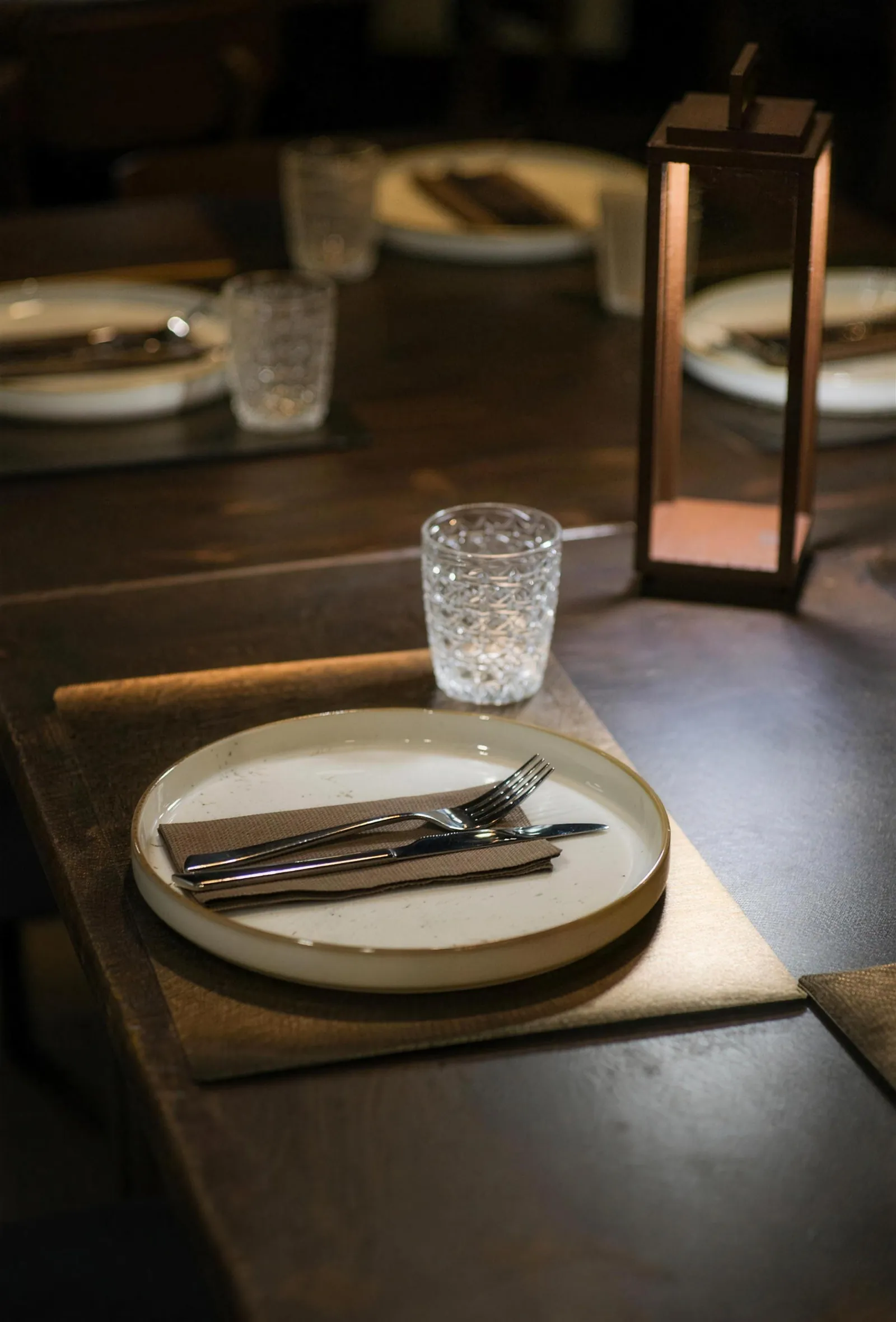 Dettaglio della mise en place: piatto in ceramica, posate, bicchiere e lanterna in rame.