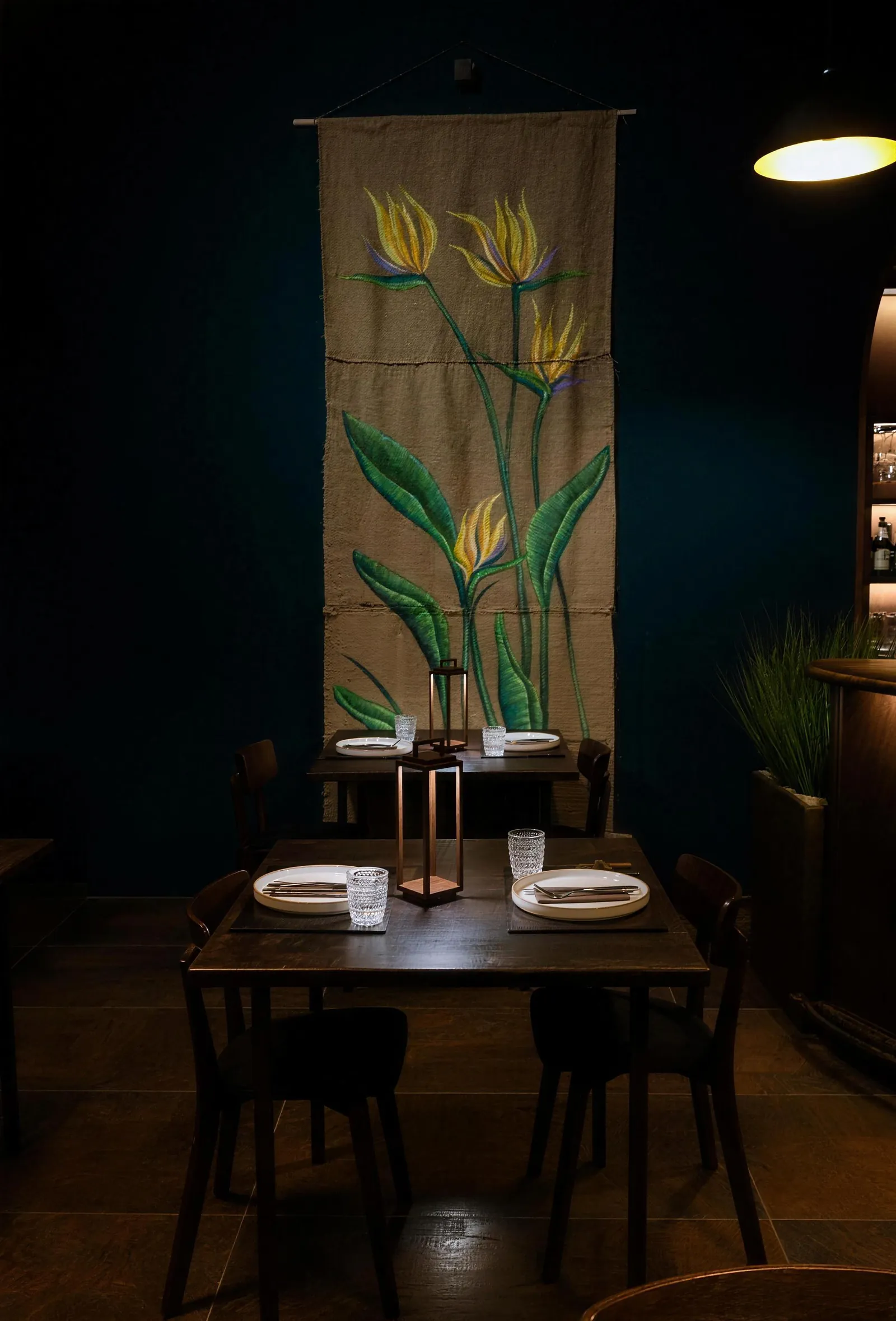 Tavolo apparecchiato sotto un arazzo dipinto con fiori gialli, in una sala blu notte.