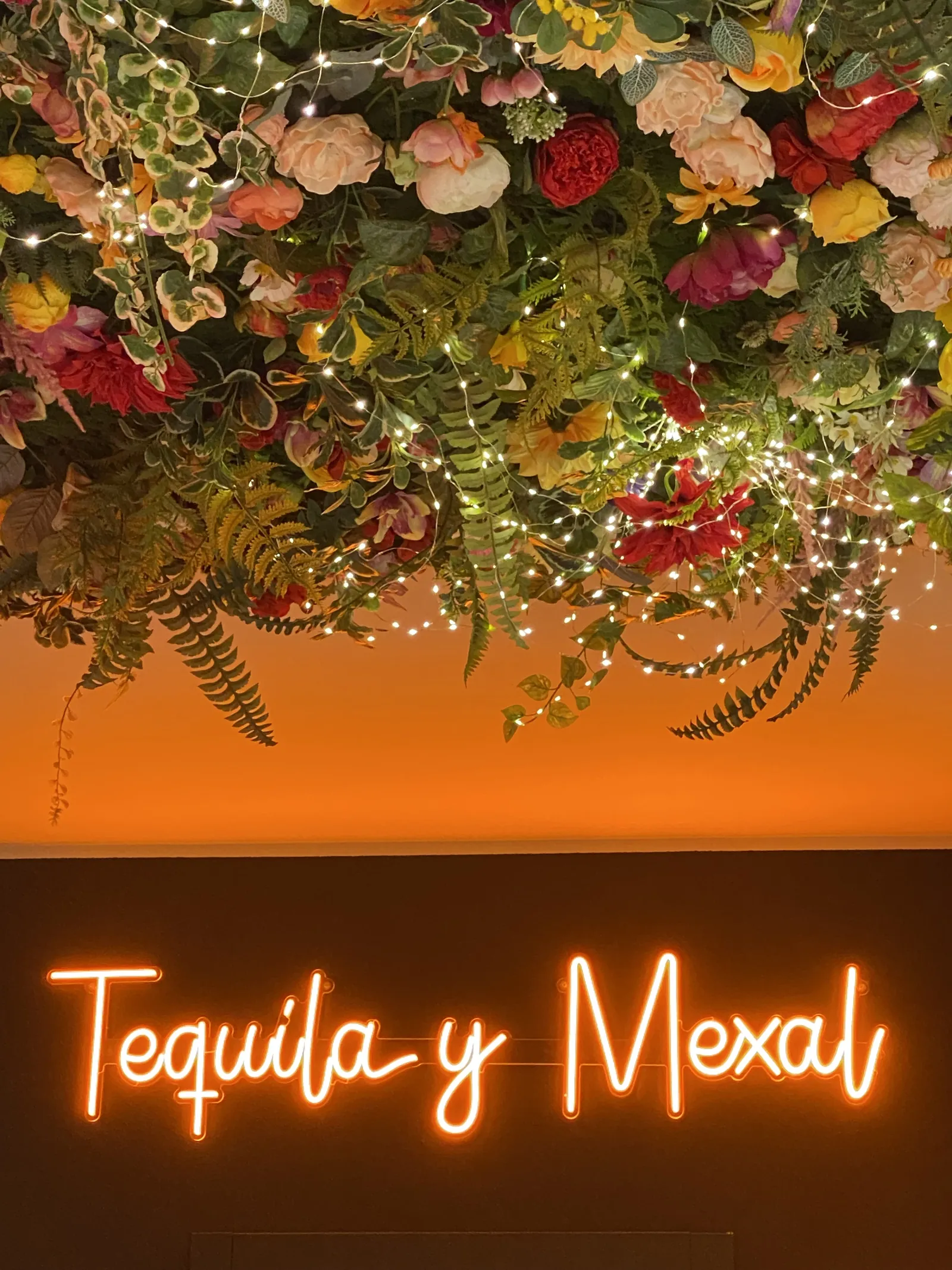 Soffitto di fiori e lucine sopra un’insegna al neon arancione con la scritta «Tequila y Mexal».