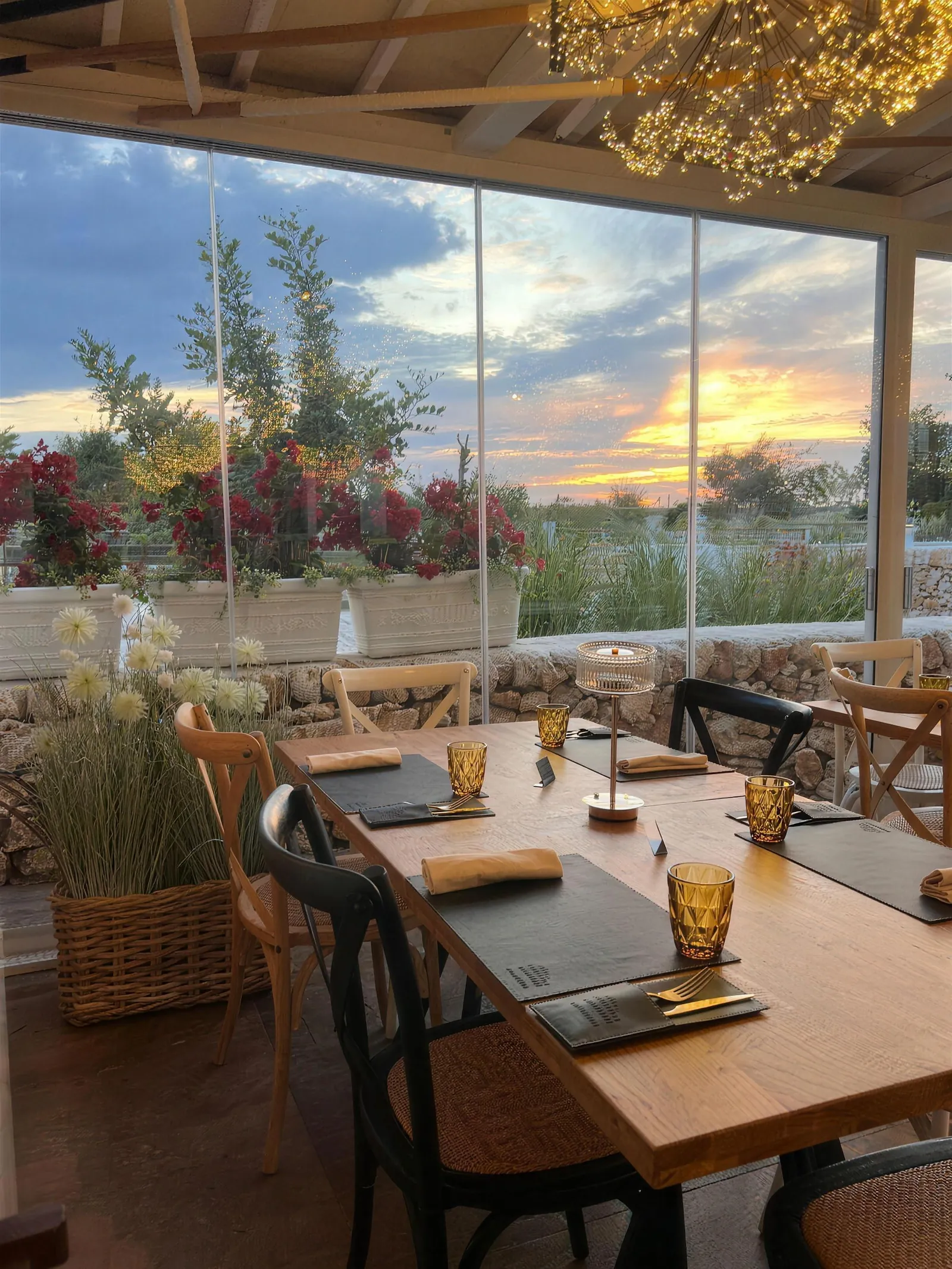 Veranda vetrata al tramonto con tavoli in legno apparecchiati e bouganville sullo sfondo.