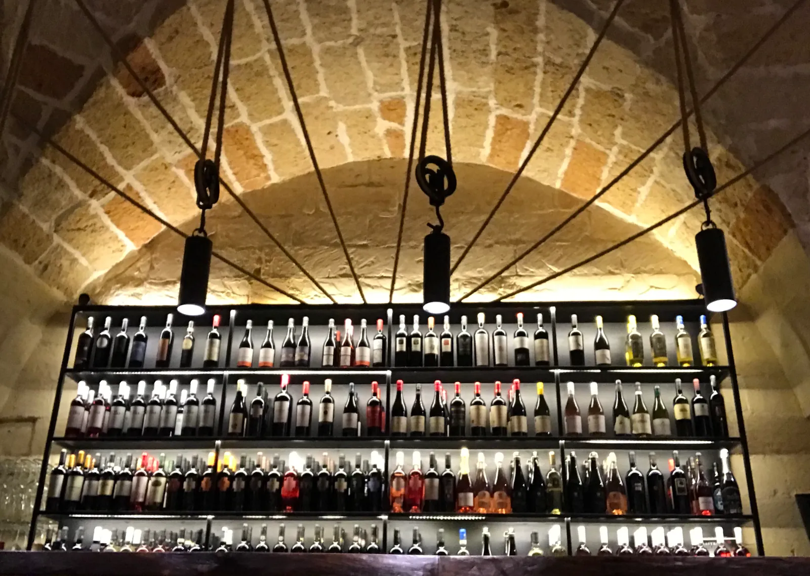 Scaffale di bottiglie di vino sospeso con tiranti sotto una volta in pietra, illuminato dal basso.